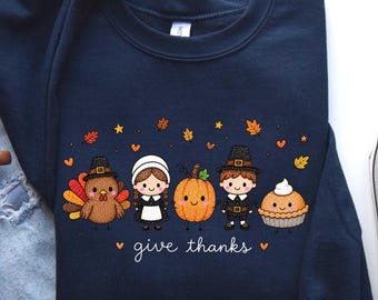 Könnte beinhalten: Ein marineblaues Sweatshirt mit Rundhalsausschnitt und einer bunten Stickerei, die einen Truthahn, einen Pilger, einen Kürbis, einen weiteren Pilger und einen Kuchen zeigt. Der Text "give thanks" ist unter den Figuren eingestickt.