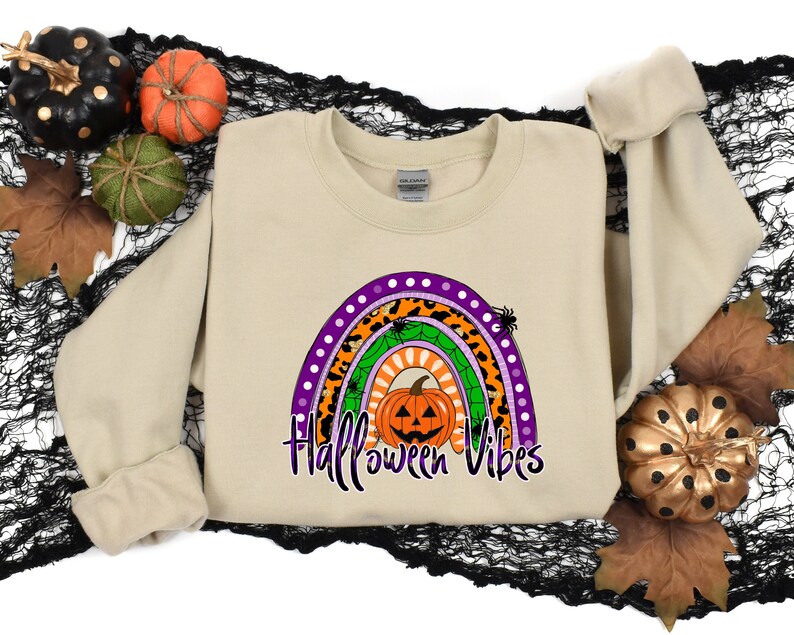 May include: A tan sweatshirt with a rainbow design featuring a pumpkin with a jack-o'-lantern face. The rainbow is made up of different patterns, including leopard print, polka dots, and spiderwebs. The text "Halloween Vibes" is printed below the rainbow.