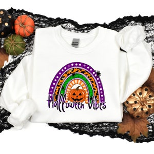 May include: White sweatshirt with a rainbow design featuring a pumpkin, spiders, and leopard print. The text "Halloween Vibes" is printed below the rainbow.