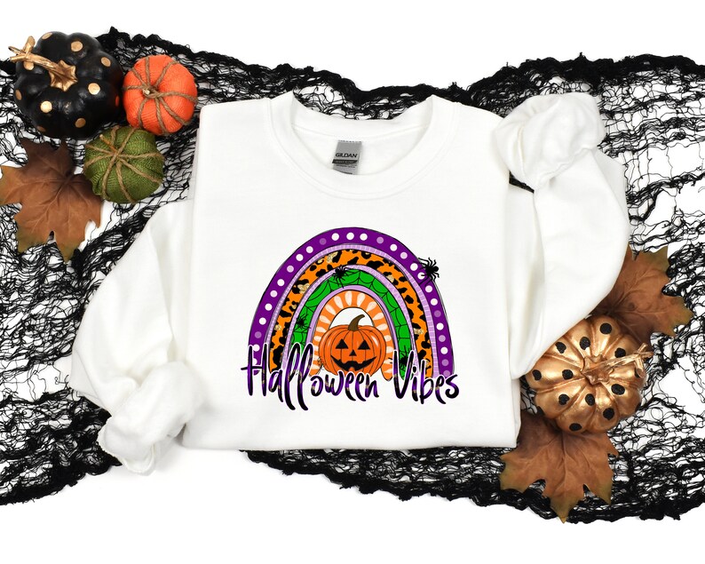 May include: White sweatshirt with a rainbow design featuring a pumpkin, spiders, and leopard print. The text "Halloween Vibes" is printed below the rainbow.