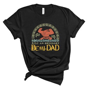 May include: Black t-shirt featuring a cartoon illustration of a muscular figure with the text "JUST AN ORDINARY DEMI-DAD" in yellow and blue. The design includes a decorative arch and water elements.