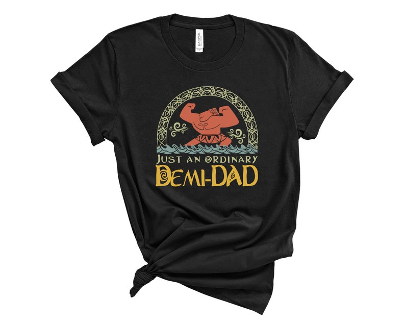 May include: Black t-shirt featuring a cartoon illustration of a muscular figure with the text "JUST AN ORDINARY DEMI-DAD" in yellow and blue. The design includes a decorative arch and water elements.