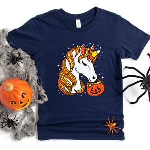 May include: A navy blue t-shirt with a white unicorn with candy corn hair holding a pumpkin filled with candy corn. The unicorn is looking at the pumpkin.
