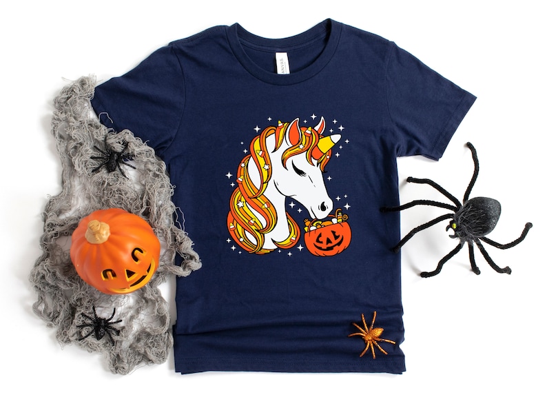 May include: A navy blue t-shirt with a white unicorn with candy corn hair holding a pumpkin filled with candy corn. The unicorn is looking at the pumpkin.