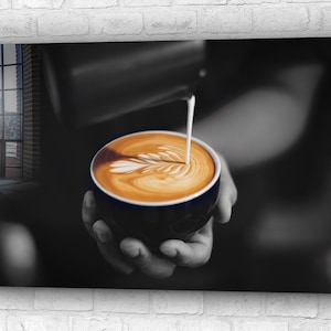 Op de afbeelding: Een close-up van een barista die melk in een kop koffie met latte art giet. De koffie zit in een donkerblauwe kop en de melk creëert een swirl-patroon bovenop de koffie.