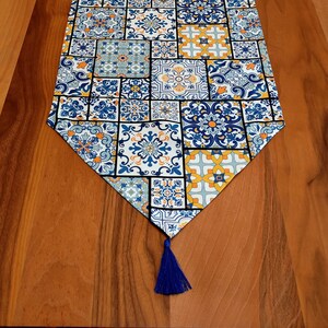 Peut inclure: Un chemin de table bleu et jaune avec un motif de carreaux géométriques. Le chemin de table a un pompon à l'extrémité.