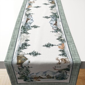 May include: A winter-themed table runner featuring a snowy forest scene with cabins, deer, and other wildlife. The runner has a light gray center with a watercolor design and a green border. The design includes evergreen trees and snow-covered mountains.