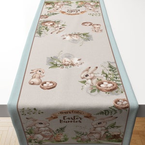 Può includere: Un runner da tavolo blu chiaro e beige con un design ad acquerello di coniglietti, nidi e uova. Il testo "Happy Easter Easter Bunnies" è stampato sul runner.