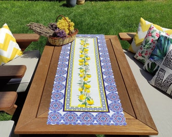 May include: A white table runner with a lemon and floral design. The runner has a blue and red border with a scalloped edge. The runner is on a wooden table with two chairs.