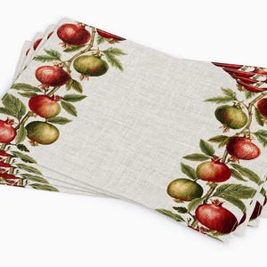 Puede incluir: Una pila de manteles individuales rectangulares con un diseño botánico. Los manteles individuales presentan un fondo beige claro con un borde de granadas rojas y hojas verdes. Las granadas varían de rojo a verde.
