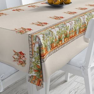 May include: A white tablecloth with a border of green plants, carrots, and brown pots. There are bunnies in red vests and a white background with a subtle pattern. The tablecloth is on a wooden table with white chairs.