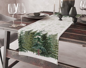 Puede incluir: Camino de mesa rectangular con una escena de bosque de árboles de hoja perenne y copos de nieve. El camino de mesa tiene un fondo blanco con copos de nieve verdes y un borde de árboles verdes. Se coloca sobre una mesa de madera oscura, creando un tema festivo.