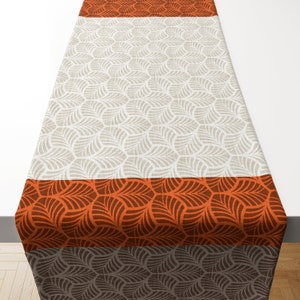 Può includere: Un runner da tavolo con un motivo geometrico di foglie in tonalità di marrone, arancione e beige. Il runner ha tre strisce di colori diversi, con la striscia superiore arancione, la striscia centrale beige e la striscia inferiore marrone.