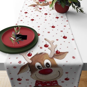 May include: White Christmas table runner featuring a cartoon reindeer with a red nose and scarf. The runner is decorated with red ornaments and snowflakes. A red plate and green plate are on the left side of the runner.