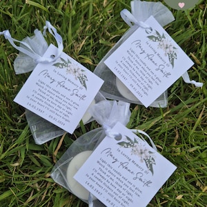 May include: Three white organza bags containing small white tea lights and a printed tag that reads "In Loving Memory of Mary Anne Smith". The tag features a floral design and includes the dates "14.03.1947 - 09.01.2024". The bags are displayed on a green grassy surface.