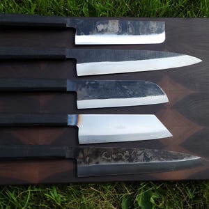 Peut inclure: Cinq couteaux de cuisine de style japonais avec des manches en bois foncé et des lames argentées sont présentés sur une planche à découper en bois foncé. Les couteaux varient en forme et en taille de lame.
