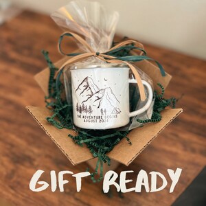 May include: A white enamel camping mug with a black mountain scene and the text "The Adventure Begins August 2024". The mug is wrapped in clear plastic and tied with a brown ribbon. The mug is sitting in a brown cardboard box with green shredded paper.