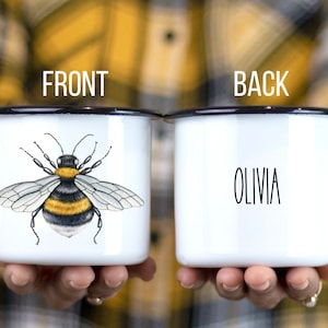 May include: White enamel mug with a black rim. The front of the mug features a black and yellow bumblebee illustration. The back of the mug has the name "Olivia" written in black.