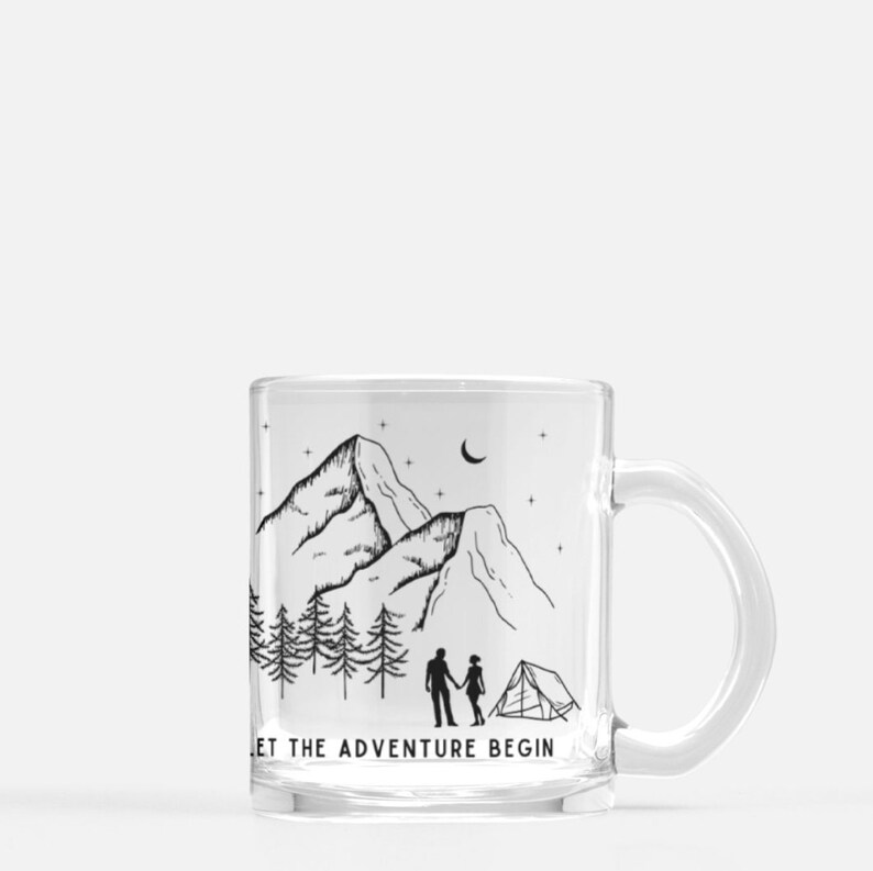 May include: A clear glass mug with a black and white illustration of a mountain range, a couple holding hands, and a tent. The text "Let the adventure begin" is printed on the bottom of the mug.
