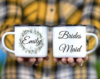 May include: Two white enamel mugs with black rims. One mug has a floral wreath design and the name "Emily" written inside. The other mug says "Brides Maid" in black cursive.