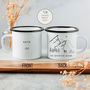 May include: Two white enamel mugs with black rims. The front mug has the names Katie and Joe printed on it. The back mug has a black and white illustration of mountains, trees, a tent, and two people standing in front of it. The text "The Adventure Begins" is printed below the illustration.