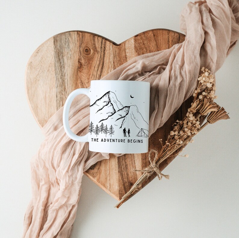 May include: White ceramic mug with a black and white illustration of mountains, trees, a couple, and a tent. The text "THE ADVENTURE BEGINS" is printed on the mug.