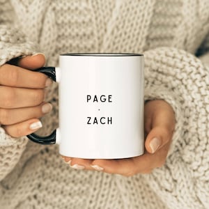 May include: White ceramic mug with a black rim and handle. The mug has the text "PAGE - ZACH" printed in black.