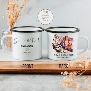May include: Two white enamel mugs with black rims. The front mug has the text "Jenna & Nich ENGAGED MAY 9, 2024" with a heart symbol. The back mug has a watercolor illustration of the Grand Canyon National Park.