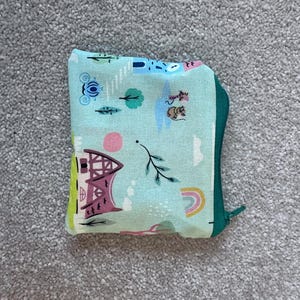 May include: A small, square, zippered pouch with a whimsical pattern. The pouch has a light blue background with colourful illustrations of houses, trees, rainbows, and other playful elements. The zipper and trim are green.