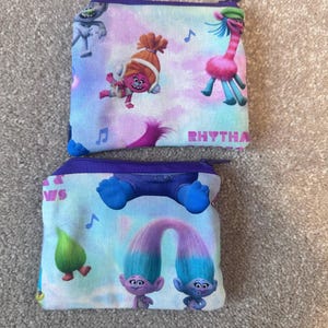 May include: Two small, rectangular zippered pouches with cartoon characters and musical notes on a light blue background. They have purple zippers and trim, with the word "RHYTHA" visible on one.
