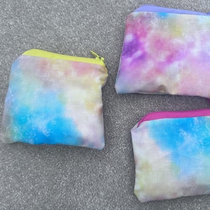 May include: Three small, rectangular pouches with zip closures. Each pouch features a tie-dye pattern in shades of blue, pink, purple, and yellow. The zips are yellow, purple, and pink.
