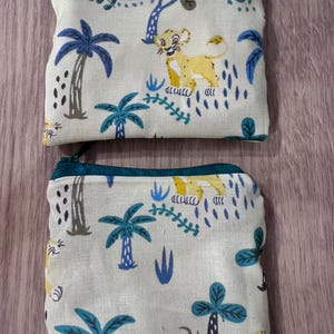 May include: Two small, rectangular pouches with a jungle-themed pattern. The pouches feature a light background with illustrations of palm trees, lions, and other jungle foliage in shades of blue, yellow, and brown. Each pouch has a zipper closure.