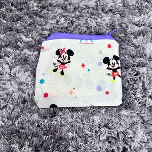 May include: A white zippered pouch with a purple zipper. The pouch has a white background with a colourful pattern of stars, dots, and Mickey Mouse and Minnie Mouse characters.