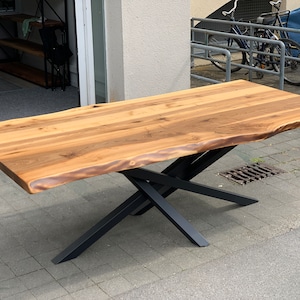 Op de afbeelding: Een grote houten tafel met een live edge blad en een zwarte metalen X-vormige basis. De tafel is gemaakt van natuurlijk hout met een lichtbruine afwerking.