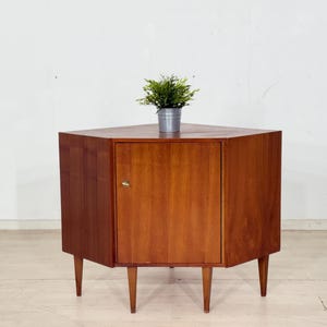 Op de afbeelding: Een houten hoekkast in mid-century modern stijl, met een driehoekige vorm en een enkele deur. De kast heeft een warme bruine kleur en taps toelopende poten. Bovenop staat een kleine plant in een pot.