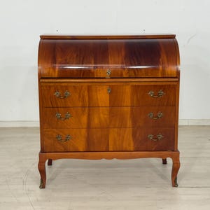 Op de afbeelding: Een vintage houten bureau met een rolluik en een warme, roodbruine afwerking. Het bureau heeft drie laden met sierlijke messing handgrepen en een gebogen bovenkant. De poten zijn sierlijk gebogen.