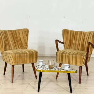 May include: Two mid-century modern armchairs with a striped pattern in shades of gold and brown. A small, triangular table with a mosaic tile top and black legs sits between the chairs. A candle holder and two cups and saucers are on the table.