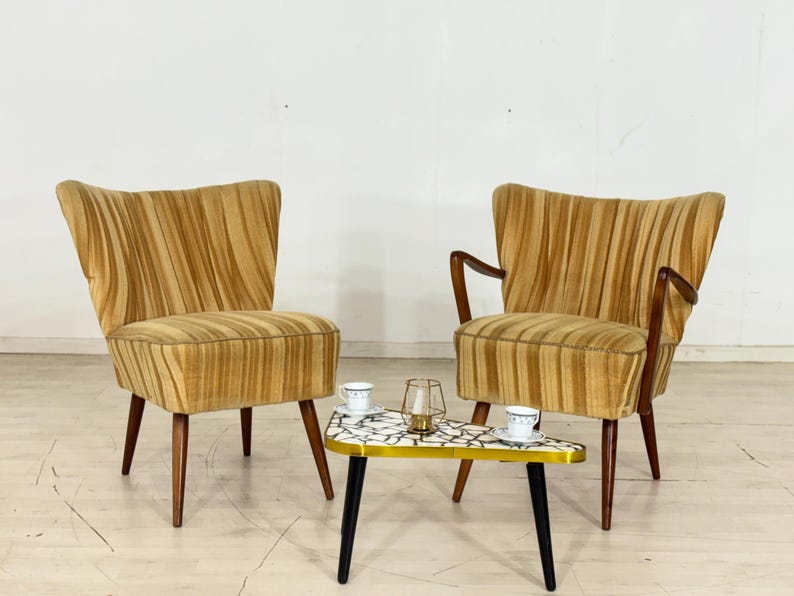 May include: Two mid-century modern armchairs with a striped pattern in shades of gold and brown. A small, triangular table with a mosaic tile top and black legs sits between the chairs. A candle holder and two cups and saucers are on the table.