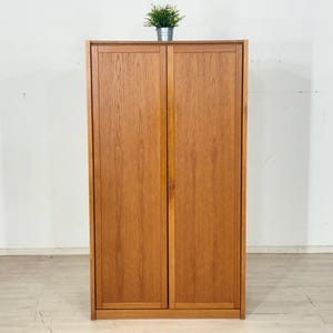 Op de afbeelding: Een houten kast met twee deuren. De kast is gemaakt van lichtbruin hout en heeft een eenvoudig, rechthoekig ontwerp. Bovenop de kast staat een kleine plant in een pot.