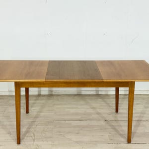 Peut inclure: Table de salle à manger rectangulaire en bois avec une finition marron clair. La table a une rallonge centrale et quatre pieds effilés. La table mesure environ 152 cm de long et 76 cm de large.