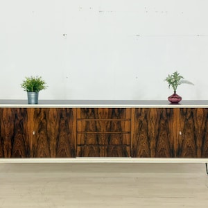 Op de afbeelding: Mid-century modern dressoir met een donkere houtnerf afwerking en drie lades. De kast heeft een zwart blad, witte rand en zwarte hairpinpoten. Twee kleine potplanten staan bovenop.