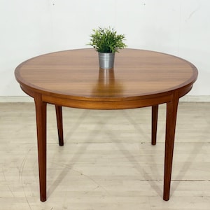 Peut inclure: Une table ronde en bois de couleur marron clair. Elle présente un bord plus foncé et quatre pieds fins et effilés. Une petite plante en pot est posée sur le dessus. La table mesure environ 100 cm de diamètre.