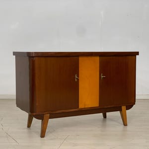 Könnte beinhalten: Ein moderner Holzschrank aus der Mitte des Jahrhunderts mit zwei Türen und einer hellholzfarbenen Platte in der Mitte. Der Schrank hat eine dunkelbraune Farbe und steht auf vier Beinen.