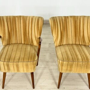 May include: Two vintage armchairs with a golden yellow, vertically striped fabric. The chairs have wooden legs and armrests. The design is mid-century modern, with a curved back and a low profile.