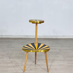 Op de afbeelding: Een plantenstandaard met twee niveaus in een modern design uit het midden van de eeuw. De standaard heeft een houten frame met drie poten en een centrale steun. De twee cirkelvormige niveaus hebben een zwart en geel zonnestraalpatroon met een gouden rand. De standaard is ongeveer 61 cm hoog.