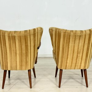 May include: Two mid-century modern armchairs with a striped, golden-yellow upholstery. The chairs have a curved back and wooden legs. The design is a classic example of vintage furniture, suitable for living rooms or lounges.