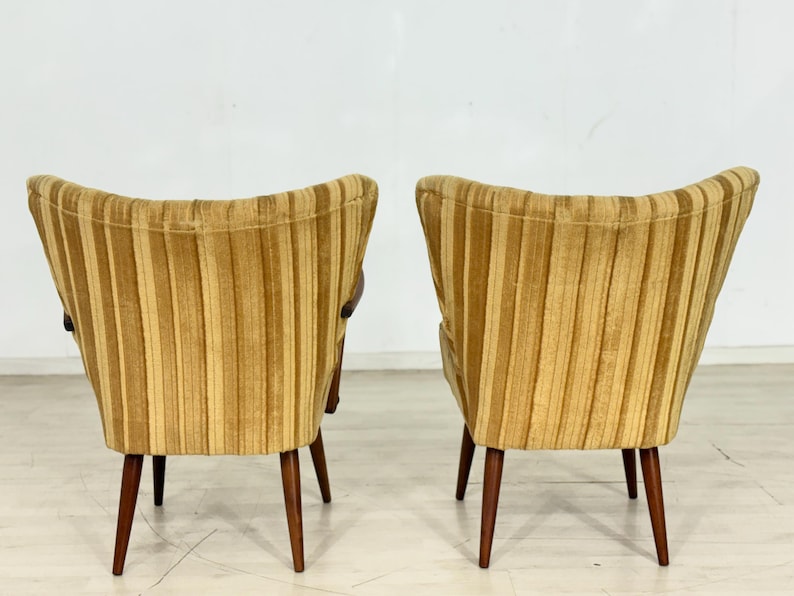May include: Two mid-century modern armchairs with a striped, golden-yellow upholstery. The chairs have a curved back and wooden legs. The design is a classic example of vintage furniture, suitable for living rooms or lounges.