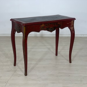 Può includere: Una scrivania in legno rosso con un piano in pelle nera e dettagli dorati decorativi. La scrivania ha un cassetto singolo e gambe curve.