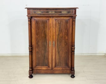 May include: A tall, rectangular wooden cabinet featuring two vertical doors with prominent wood grain patterns. The piece includes a top drawer with an ornate metal keyhole plate and decorative columns on both sides, supported by four feet.