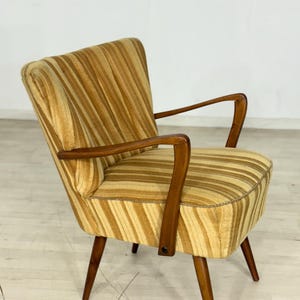 May include: A vintage armchair with a striped pattern in shades of gold and brown. The chair features wooden armrests and legs, showcasing a mid-century modern design. The seat and back are upholstered with a textured fabric.